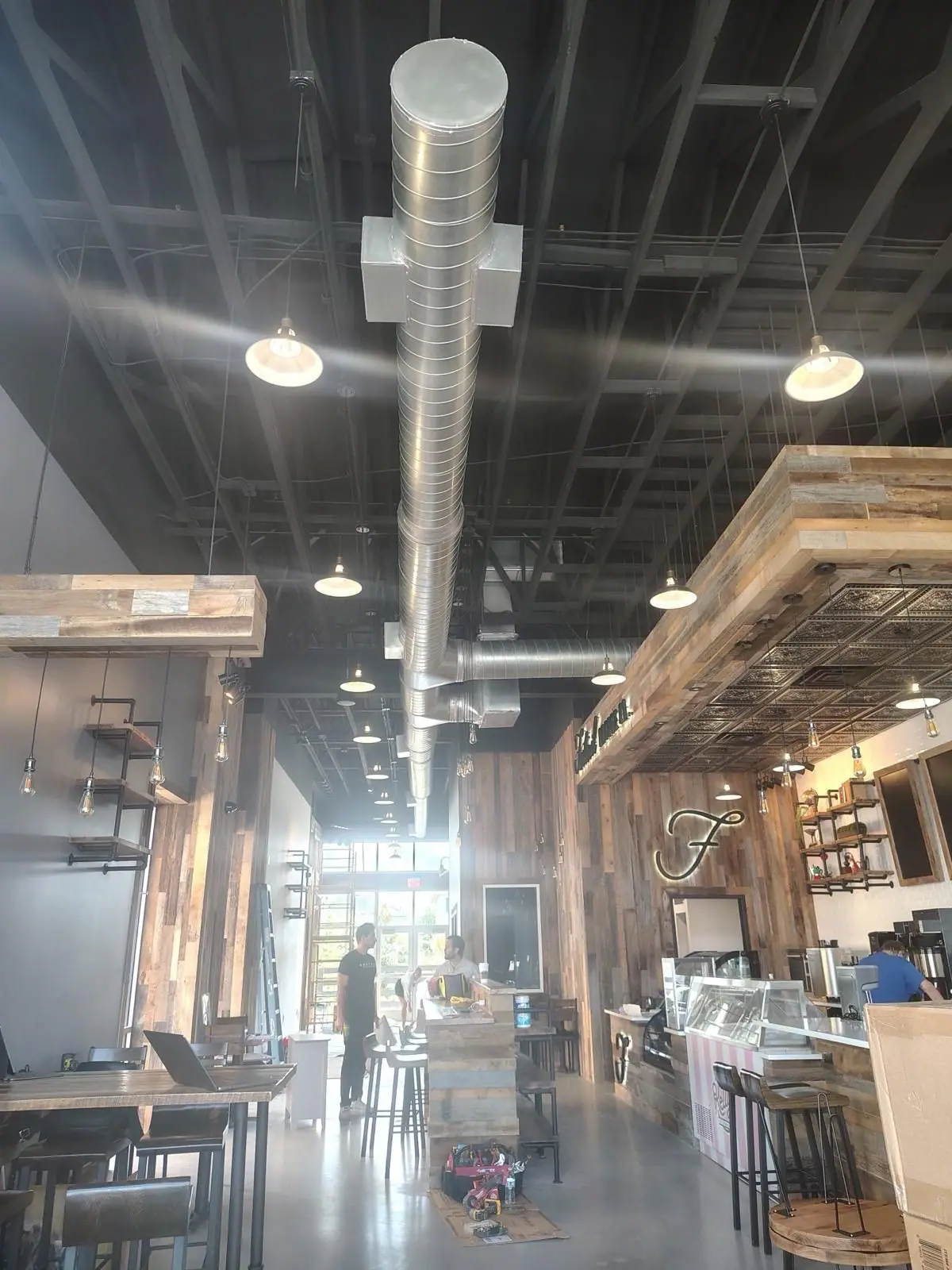 Long run of exposed spiral ductwork above a commercial interior with pendant lights and wood cladding