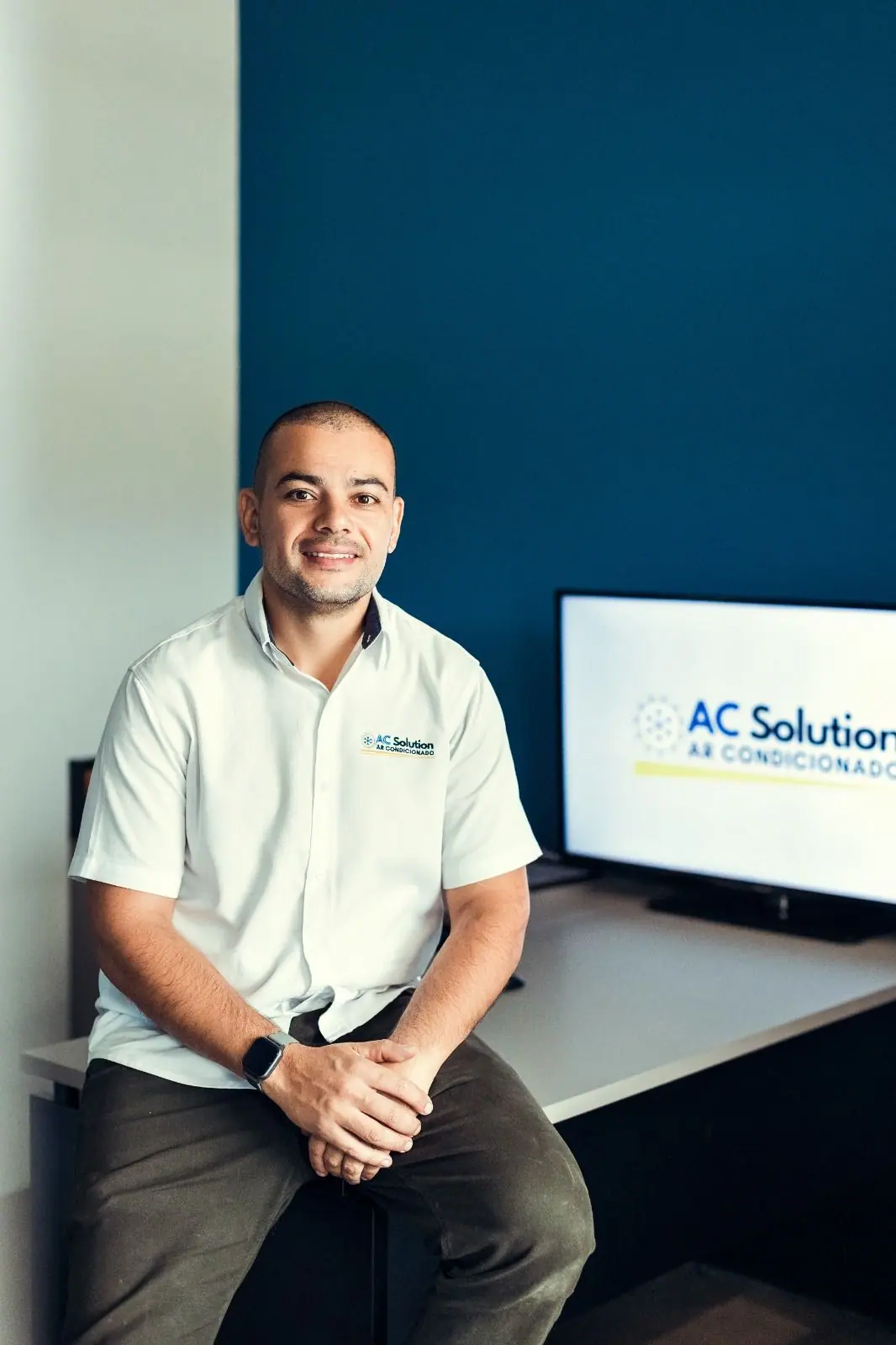 Smiling founder seated at a desk in a white company shirt, with a company logo on the screen behind